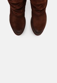 Bottes en suede marron à talon slouch avec bouts pointus et bords surpiqués vues de dessus sur un fond clair.