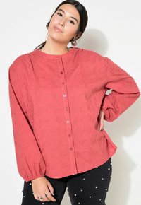 Camicia rossa con bottoni, con un motivo testurizzato, maniche lunghe e scollo rotondo. Abbinata a pantaloni neri con dettagli bianchi.