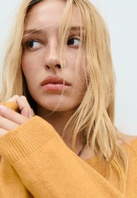 Blondýnka s volnými vlasy v hořčicovém svetru se dívá do strany, ruce blízko hrudníku, na jednobarevném světlém pozadí.