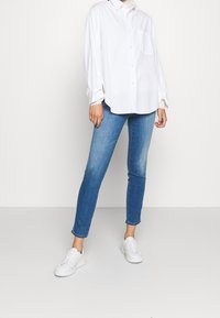 Camisa blanca de botones con mangas largas, que presenta un bolsillo en el pecho. Combinada con jeans ajustados de color azul claro y zapatillas blancas. Diseño simple y clásico.