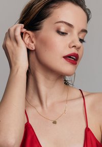 Złota naszyjnik w kształcie serca na delikatnym łańcuszku, z małą perłą jako akcentem. Modelka ma na sobie czerwoną satynową bluzkę i mokre włosy.