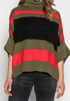 Kobieta w luźnym swetrze z krótkim rękawem, z szerokimi poziomymi paskami w kolorach oliwkowej zieleni, czerni i czerwieni, w połączeniu z ciemnymi jeansami.