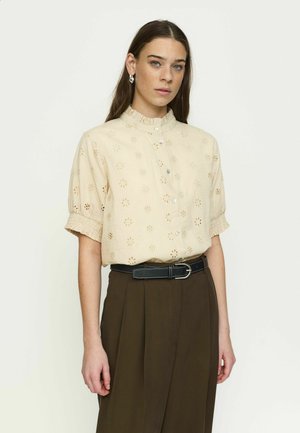 Noorpruunide pikkade juustega noor naine kannab beežikat silmikkoega pluusi ja tumedat pruuni lipsutatud pükstega koos musta vööga, seisab heleda tausta ees.