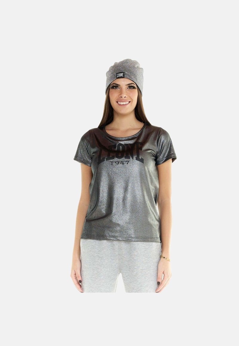 Femme portant un T-shirt gris foncé brillant avec le texte « LEONE 1947 » et un bonnet gris clair, debout sur un fond blanc.