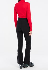 Eine Frau trägt schwarze Skihosen, ein rotes Langarmoberteil und schwarze Skischuhe. Sie steht mit dem Rücken zu einem weißen Hintergrund.