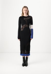 Svart stickad klänning med nätmönster, långa ärmar och blå detaljer vid ärmsluten och fållen. Har en figurnära silhuett med utskärningar.