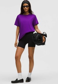 Violeta īsām piedurknēm T-kreklā, melnās augstās velosipēdu šortre, melnā soma, melnie loafers un baltas zeķes. Vienkāršs, neformāls dizains.