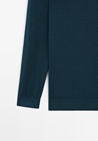 Manche d’un pull en maille bleu sarcelle foncé à manches longues avec poignets côtelés sur fond blanc uni.