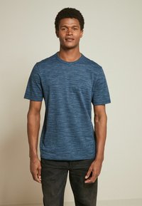 Tricou albastru cu mânecă scurtă, dintr-un material texturat și cu pattern. Design cu gât rotund. Model purtând blugi închisi la culoare. Croială simplă, casual.