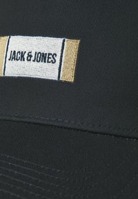 Zwarte stoffen pet met een genaaide rechthoekige patch waarop "JACK & JONES" staat in zwarte tekst op een witte en beige achtergrond.
