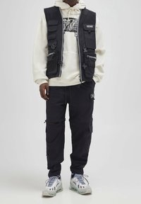 Czarna kamizelka użytkowa z wieloma kieszeniami na białej bluzie z kapturem, w połączeniu z czarnymi spodniami cargo i szarymi tenisówkami. Miękka tekstura, funkcjonalny design.