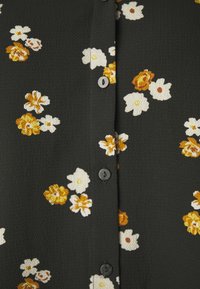 Svart blus i tyg med blommönster i vita och gula nyanser. Har knappar ned längs framsidan och en strukturerad yta.