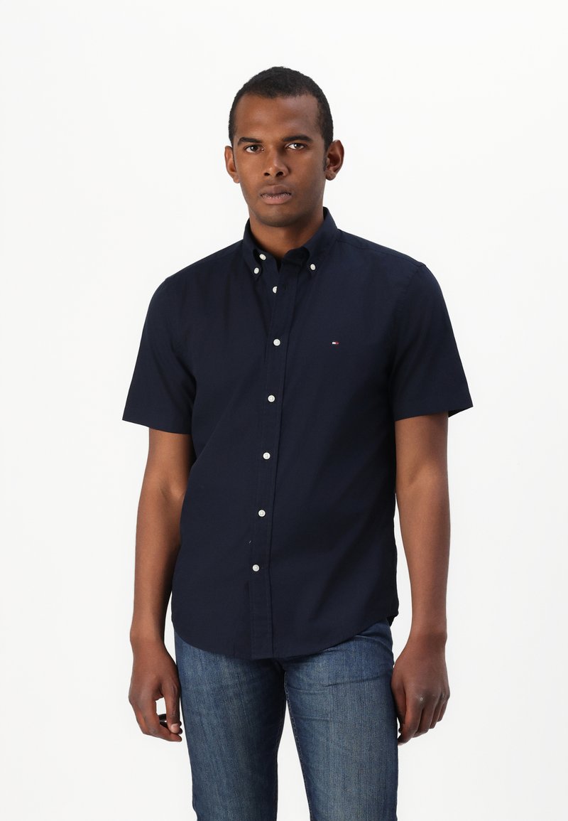 Camicia a maniche corte blu navy con bottoni bianchi e un piccolo logo. Jeans in denim con una silhouette aderente. Tessuto dalla consistenza liscia.
