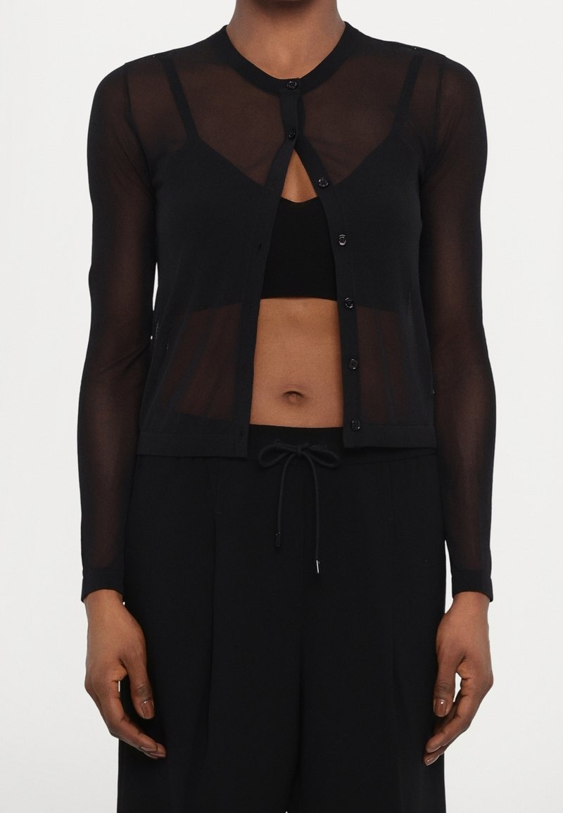 Cardigan court noir transparent à manches longues avec boutons, porté sur un crop top noir, associé à un pantalon noir à cordon.