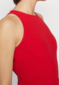 Top sans manches rouge en tissu lisse, avec un col haut et une coupe ajustée. Des détails de couture visibles mettent en valeur la structure.