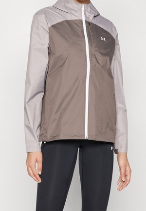 Personne portant une veste zippée bicolore grise avec capuche et des leggings noirs, debout, les bras détendus le long du corps.