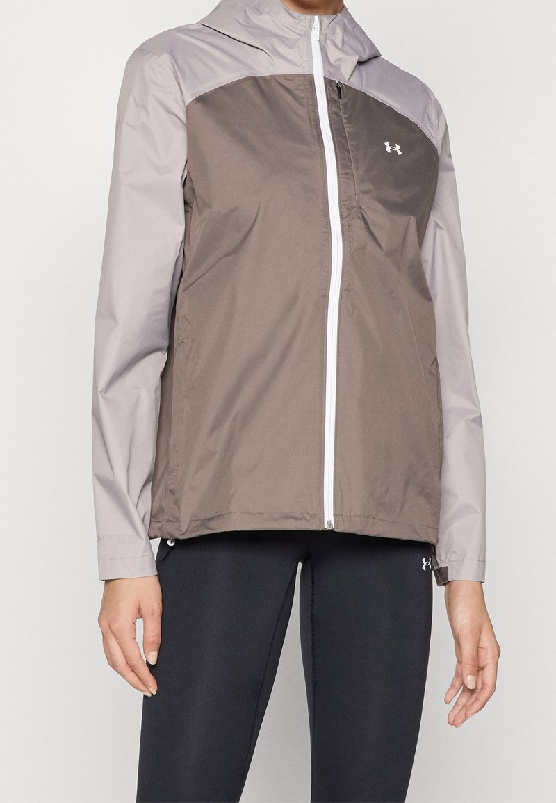Personne portant une veste zippée bicolore grise avec capuche et des leggings noirs, debout, les bras détendus le long du corps.