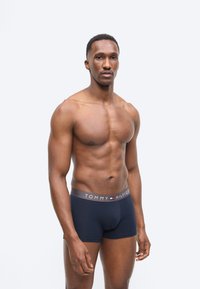 Μυώδης άνδρας που στέκεται μπροστά σε απλό φόντο, φορώντας σκούρο μπλε μποξεράκια Tommy Hilfiger, κοιτώντας μπροστά με ουδέτερη έκφραση.