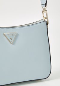 Sac à main en cuir synthétique bleu clair avec un logo triangulaire argenté, fermeture éclair sur le dessus et détail de bordure noire. Texture lisse, design moderne.