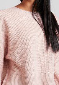 Hellrosa gestrickter Pullover mit geripptem Muster und Rundhalsausschnitt. Der Stoff wirkt weich und leicht locker sitzend.