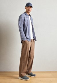 Chemise bleue à carreaux, t-shirt blanc, pantalon beige large, chaussures grises et casquette grise. Tenue décontractée avec des détails à boutons et une coupe décontractée.
