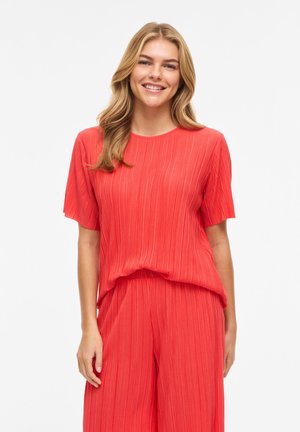 Femme souriante aux longs cheveux blonds portant un haut plissé rouge vif à manches courtes et un pantalon large assorti sur un fond blanc.