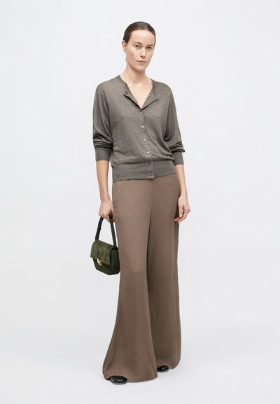 Nő szürke gombos kardigánban és bő szárú barna nadrágban, kezében egy kis sötétzöld kézitáskát tart, egyszínű fehér háttér előtt áll.