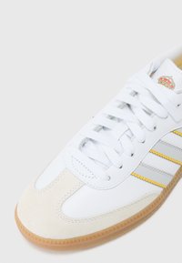 Chaussure de sport blanche en matériaux de daim et de cuir, avec des rayures grises, des accents jaunes et une semelle en gomme au motif texturé.