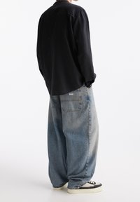 Persoon in een zwart langmouw shirt en een losse lichtblauwe spijkerbroek met wijde pijpen, gekleed in zwart-witte sneakers, staande met de rug naar de kijker gekeerd.