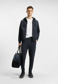 Laivaston sininen vetoketjullinen huppari ja siihen sopivat joggerit sileää kangasta, yhdistetty valkoiseen t-paitaan. Pitelee kaksiväristä urheilukassia ja jalassaan mustat lenkkarit.