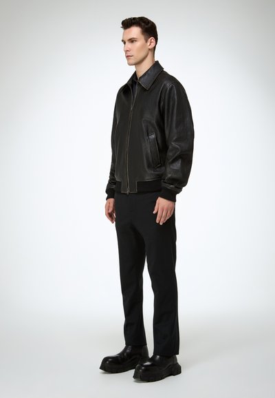 Hombre con chaqueta de cuero negra, pantalones negros y botas negras gruesas, de pie contra un fondo claro y liso, mirando hacia la izquierda.