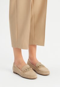 Beige mokasiner i mocka med platt sula, som har en dekorativ kedjedetalj på framsidan och en rundad tåsdesign.