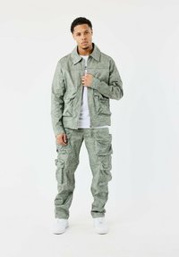 Lichtgroene, gestructureerde jas met grote zakken, gecombineerd met bijpassende cargobroek. De outfit heeft een ritssluiting en witte sneakers.
