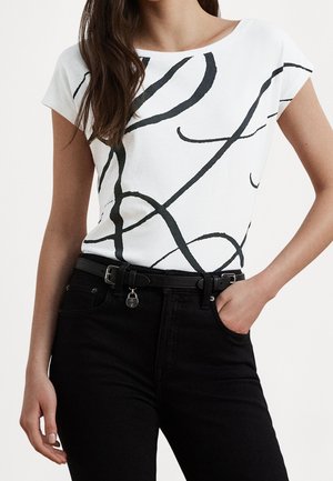 Haut blanc à manches courtes avec des lignes noires abstraites, porté avec un pantalon noir et une ceinture noire avec un petit pendentif cadenas.