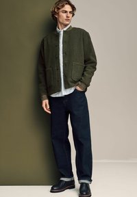 Green textured jacket with two front pockets, layered over a blue and white striped shirt, paired with dark navy jeans and black shoes.