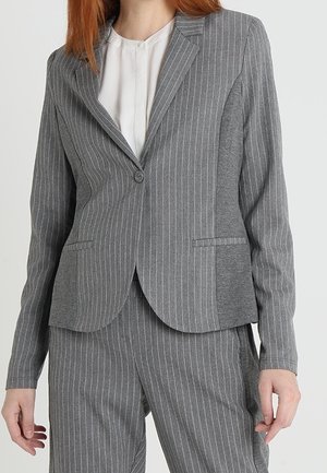 Femme portant un blazer gris à fines rayures avec un bouton et un pantalon assorti à fines rayures sur une blouse blanche.