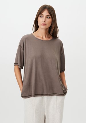 Mujer con cabello castaño, que lleva una camisa suelta de manga corta color topo y pantalones beige claros, de pie con las manos en los bolsillos, mirando hacia un lado.