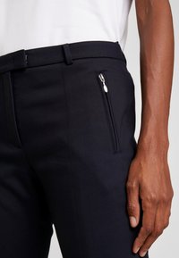 Pantalons bleu marine avec une coupe slim, en tissu lisse, dotés d'une poche zippée à l'avant, associés à un haut blanc léger.