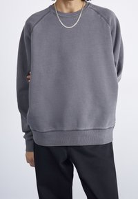 Sudadera gris con cuello redondo, puños acanalados y un ajuste ligeramente holgado. Textura suave, con mangas raglán y un diseño simple.