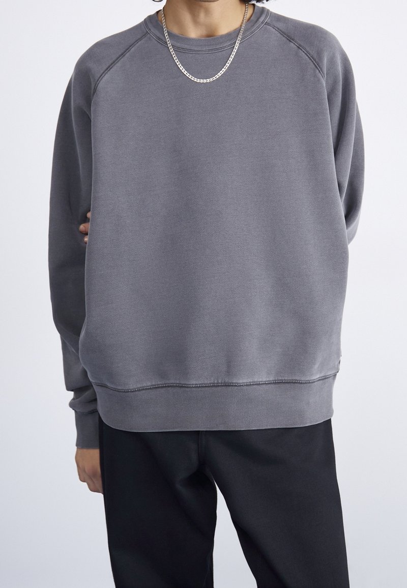 Sudadera gris con cuello redondo, puños acanalados y un ajuste ligeramente holgado. Textura suave, con mangas raglán y un diseño simple.