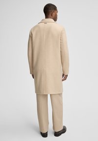 Cappotto di lana beige con colletto singolo, design dritto e spacchetto sul retro. Abbinato a pantaloni leggeri plissettati e calzature nere.