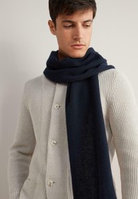 Ciemnoniebieski szalik z miękkiego dzianiny, zarzucony na szyję, w połączeniu z jasnobeżowym kardiganem w prążki, wyposażonym w duże guziki.