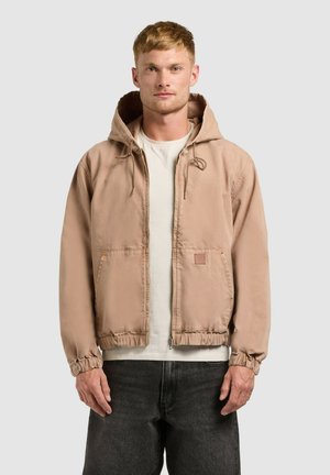 Jonge man die recht vooruit kijkt, gekleed in een beige jack met rits en capuchon, een wit T-shirt en donkere spijkerbroek, tegen een effen achtergrond.