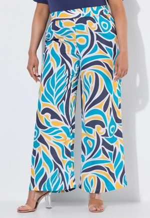 Weite Hosen mit einem abstrakten, bunten Muster in Blau, Gelb und Schwarz, kombiniert mit transparenten Sandalen mit hohen Absätzen.