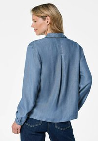 Blaue Jeanshemd mit langen Ärmeln, einer Rückenfalte und einem klassischen Kragen. Weiche Textur, abgerundeter Saum und geknöpfte Manschettendetails.