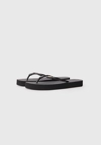 Schwarze Flip-Flops mit einer strukturierten Sohle und dünnen Riemen, die ein kleines metallisches Logo-Detail am Riemen aufweisen. Einfaches, flaches Design.