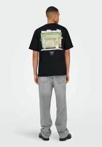 Sort t-shirt med korte Ærmer, der har en farverig grafik af en butik på ryggen. Kombineret med lysegrå, løstsiddende jeans og sorte sko.