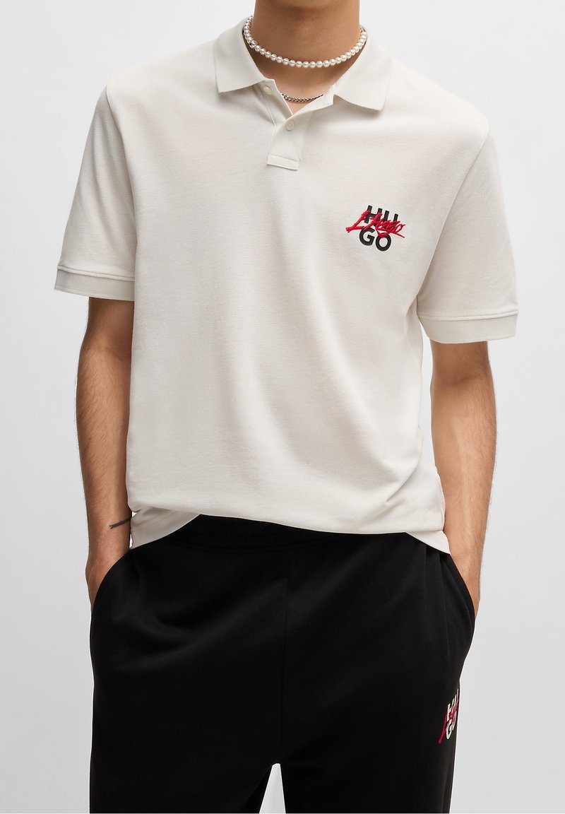 Homme portant un polo écru avec un logo rouge et noir sur la poitrine et un pantalon noir avec le même logo, mains dans les poches, collier de perles blanches.