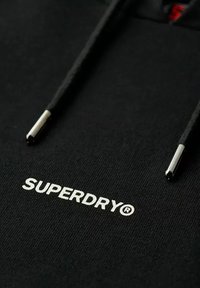 Zwarte hoodie met trekkoorden, voorzien van een gestructureerde afwerking. De merknaam "SUPERDRY" is in het wit geborduurd, vergezeld van zilveren aglets.