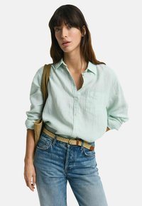Lichtgroen linnen overhemd met knopen en een borstzak, gecombineerd met blauwe denim jeans en een geweven beige riem. Schoudertas zichtbaar.